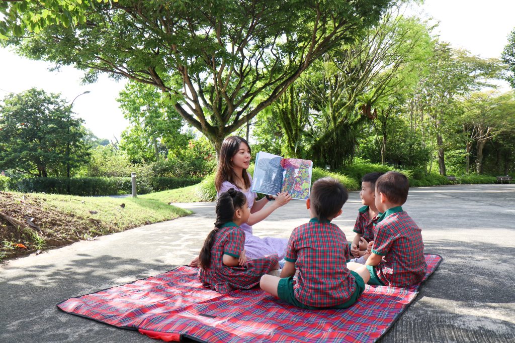 EtonHouse Riyadh - A teacher teaching showing a book to preschoolers sitting near a garden road
