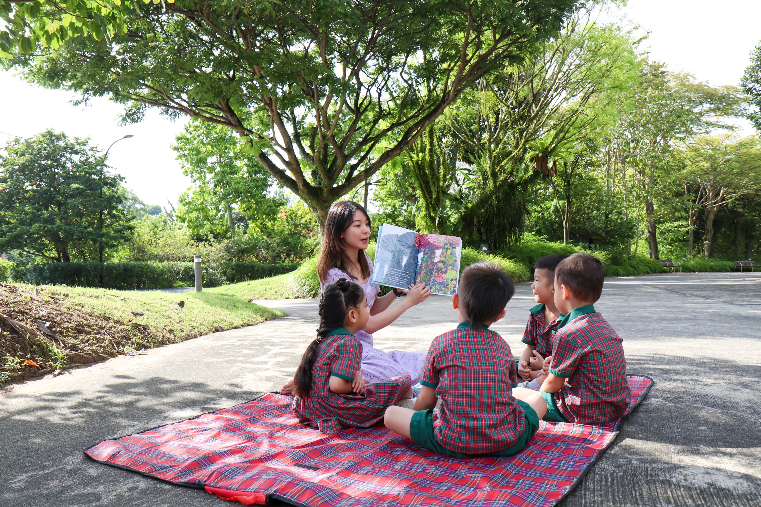 EtonHouse Riyadh - A teacher teaching showing a book to preschoolers sitting near a garden road