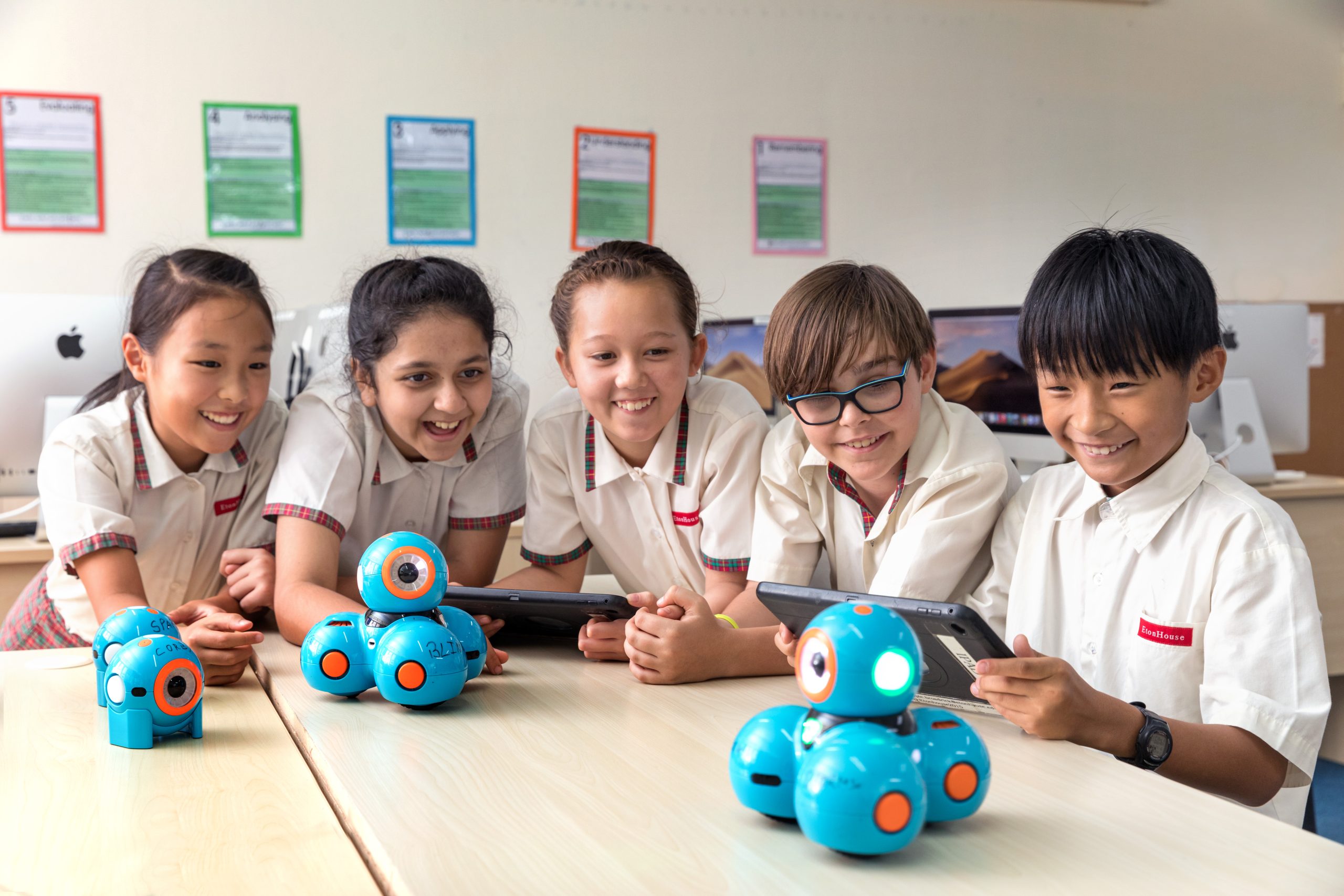 EtonHouse Riyadh - Five primary-school students gathered around blue educational robots and tablets, smiling and engaged in collaborative learning.