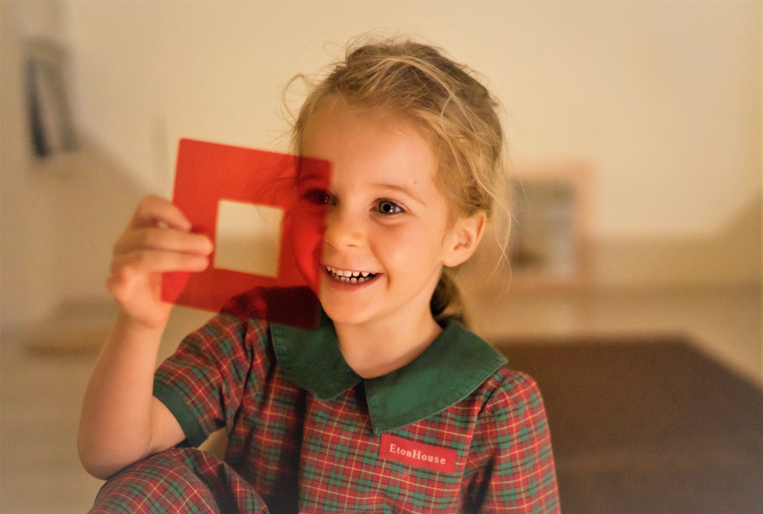 EtonHouse Riyadh - A smiling preschool-aged child holding up a red square cut-out, wearing a checkered EtonHouse uniform.